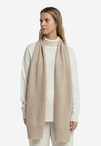Écharpe en tricot beige fabriquée en laine douce, avec une texture lisse et des bords côtelés, portée sur un pull col roulé blanc.