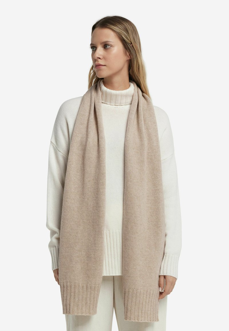 Écharpe en tricot beige fabriquée en laine douce, avec une texture lisse et des bords côtelés, portée sur un pull col roulé blanc.