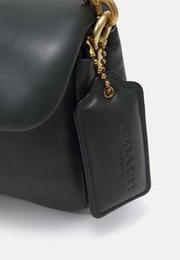 Vihreä nahkainen käsilaukku, jossa on kultainen ketjusomiste ja musta nahkainen merkki, johon on painettu "COACH NEW YORK." Sileä pinta, jossa hillitty kiilto.