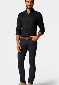 Camicia nera con bottoni e maniche lunghe, abbinata a pantaloni slim-fit navy e una cintura intrecciata marrone. Sneakers beige completano il look.