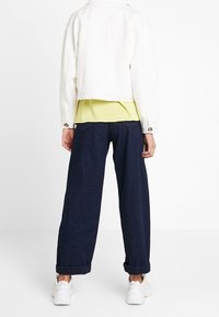 Hvid cropped jakke over en gul skjorte, parret med mørkeblå vide jeans. Har opsmøgede ærmer og hvide sneakers. Simpelt design.