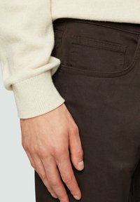 Braune Baumwollhosen mit einer Gesäßtasche, kombiniert mit einem cremefarbenen Strickpullover. Der Pullover hat ein geripptes Bündchen und umgeschlagene Ärmel.