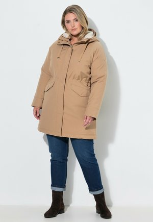 Kvinde iført en beige hættejakke til vinteren over blå jeans og mørke ankelstøvler, stående mod en ensfarvet lys baggrund.