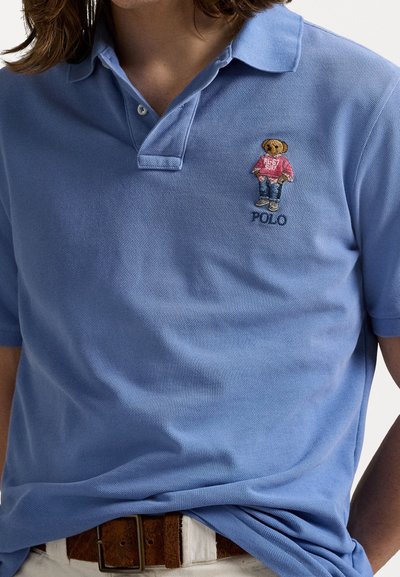 Light blue short-sleeve polo shirt made of textured fabric, featuring an embroidered bear design and "Polo" text on the chest.