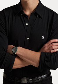 Zwart polohemd met lange mouwen, een spitse kraag, witte knopen en een klein wit logo. Model draagt een zilveren horloge met een zwarte band.