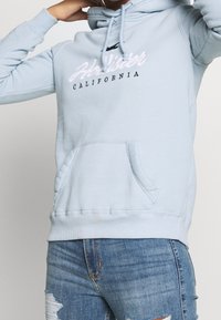 Felpa con cappuccio azzurro chiaro con tasca frontale, cappuccio con coulisse e testo ricamato "Hollister California" in bianco e nero. Tessuto morbido in misto cotone.
