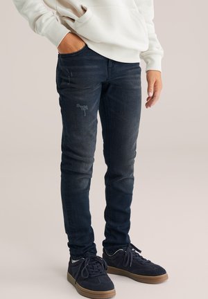 Persoon draagt donkerblauwe slim-fit jeans met lichte slijtage, zwarte sneakers met bruine zolen en een witte hoodie, met één hand in de zak.