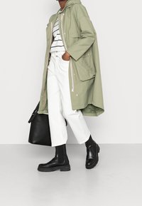 Jasnozielona parka z kapturem zapinana na guziki, z kieszeniami, biała koszula w paski, białe spodnie o szerokich nogawkach oraz czarne botki za kostkę, zestawione z czarną torbą.