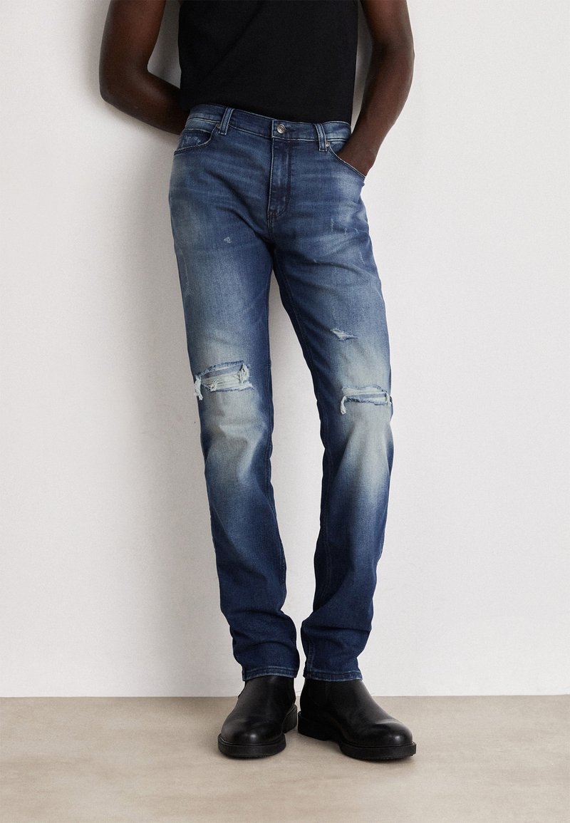 Homme portant un jean bleu délavé déchiré et des bottines noires, debout les mains dans les poches devant un mur uni.