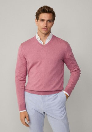 Helles rosa V-Ausschnitt-Pullover aus weichem Material mit langen Ärmeln und taillierter Passform. Kombiniert mit hellblauen gestreiften Hosen.