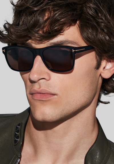 Gafas de sol rectangulares negras con lentes oscuros, acentos dorados en las varillas y un acabado brillante. El modelo tiene el cabello despeinado y lleva una chaqueta de cuero.