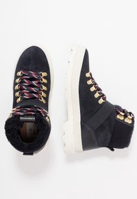 Black suede ankle boots with a white chunky sole, gold eyelets, red and blue laces, and a fuzzy interior lining.