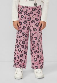 Bambina che indossa pantaloni larghi rosa con stampa floreale scura e sneakers bianche, in piedi contro uno sfondo semplice.