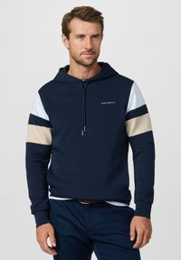 Muž oblečený v námornícky modrej mikine s kapucňou s béžovými a bielymi pruhmi na rukávoch a logom "Hackett", stojaci pred jednoduchým svetlým pozadím.