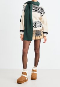 Personne portant un pull à motifs beige et noir, une écharpe verte, une mini jupe tie-dye, des collants noirs transparents et des bottines à fourrure beige.