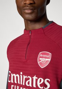 Maillot d'entraînement Arsenal couleur bordeaux avec un col zippé, présentant un design texturé et des accents blancs pour le logo et le sponsor, mettant en valeur l'identité de l'équipe.