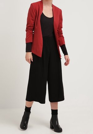 Femme portant un blazer rouge sur un haut noir, des culottes noires à jambes larges et des bottines noires à talons, debout sur un fond uni.