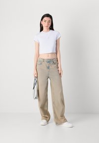 Een jonge vrouw staat zelfverzekerd, gekleed in een cropped wit t-shirt en losse lichte jeans, terwijl ze een zilveren handtas vasthoudt. Eenvoudige achtergrond.