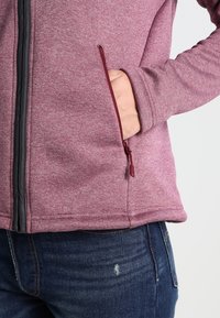 Polarowa kurtka w melanżowym burgundowym kolorze z zamykaną na zamek kieszenią boczną i kontrastowym ciemnoszarym zamkiem. Miękka tekstura i luźny krój.