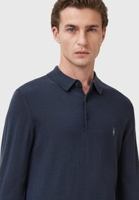 Marineblauwe polo met lange mouwen, gemaakt van zacht materiaal. Voorzien van een kraag, knoopsluiting en een klein logo in lichte draad op de borst.