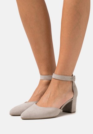 Pieds portant des chaussures à talons blocs en suède gris clair avec des brides de cheville et des boucles en argent sur un fond blanc.