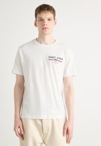 Hvid t-shirt med korte ærmer lavet af bomuld. Har "GANT 1949" logo i marineblå og rød på venstre bryst. Glat tekstur, klassisk pasform.