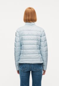 Άτομο με ίσια κόκκινα μαλλιά που φορά ανοιχτό μπλε γιλέκο με καπιτονέ και μπλε τζιν, φαίνεται από πίσω σε λευκό φόντο.