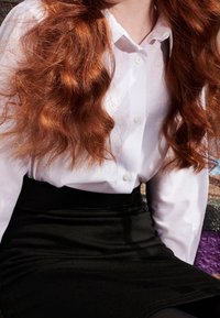 Chemise blanche à boutons avec col, associée à une jupe noire ajustée. Les cheveux sont longs, ondulés et châtain rougeâtre.