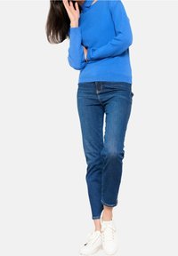 Pull en tricot bleu à col en V, associé à un jean denim délavé moyen et des baskets blanches. Accent sur la texture et la coupe décontractée.