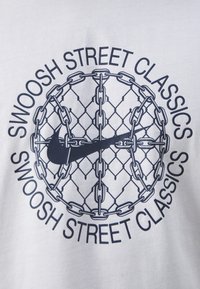 Witte T-shirt met een cirkelvormige graphic met kettingen en een hekpatroon, waarop het Nike-logo en "SWOOSH STREET CLASSICS" in vetgedrukte letters zijn weergegeven.