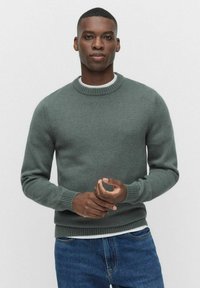 Tumma vihreä neulepaita, jossa on pyöreä kaula-aukko ja ribbireunukset, yhdistetty valkoiseen sisäkerrokseen ja sinisiin farkkuihin. Silkkinen pinta.