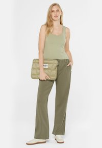 Olivengrünes Tanktop kombiniert mit einer passenden weit geschnittenen Hose, hält eine gesteppten, hellgrünen Clutch mit einem schwarzen Etikett. Weiße Sneaker.