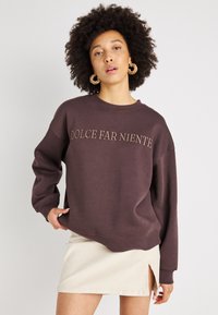 Sweatshirt castanho escuro com "DOLCE FAR NIENTE" bordado em castanho claro. Tem um corte solto, gola canelada e ombros caídos.