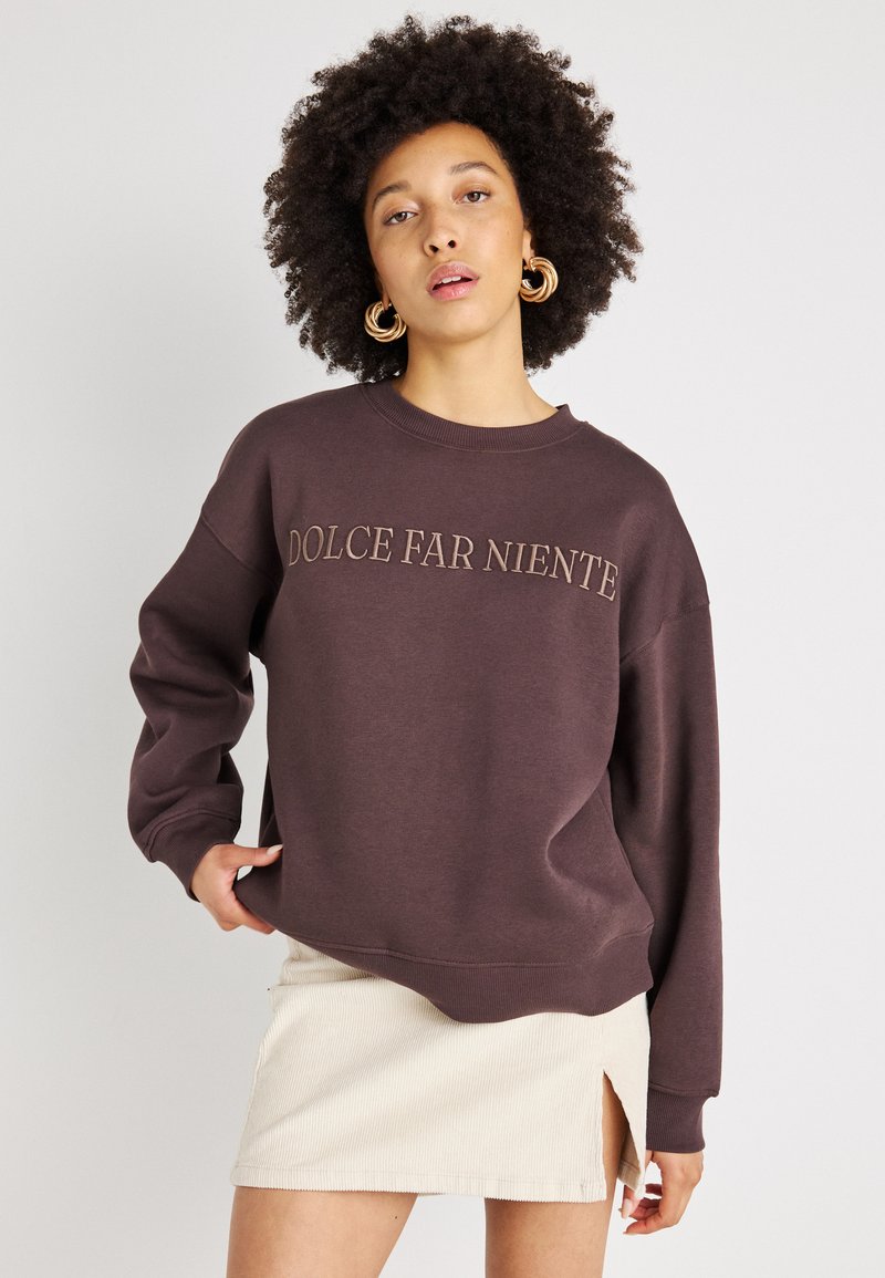 Sweatshirt castanho escuro com "DOLCE FAR NIENTE" bordado em castanho claro. Tem um corte solto, gola canelada e ombros caídos.