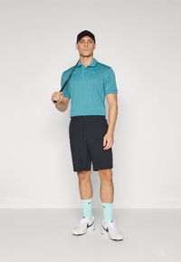 Turkizna polo majica z diskretnim vzorcem, črne kratke hlače in bele športne copate s sivo-naglasitvijo. Drži golf palico, nosi črno kapo.