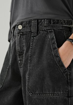 Person iført sorte denimbukser med store forlommer og et sort armbånd, hånden delvist i lommen, mod en neutral baggrund.