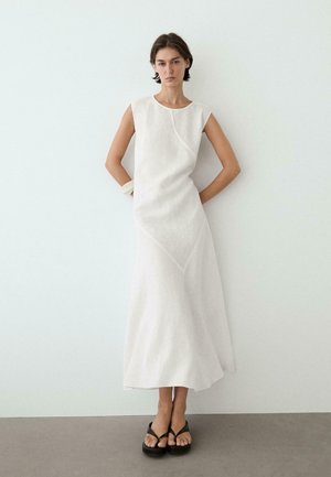 Frau steht an einer weißen Wand, trägt ein ärmelloses weißes Midikleid mit asymmetrischen Nähten, schwarze Plateausandalen und ein weißes Armband.