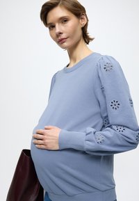 Svijetloplavi pulover s naboranim rukavima i cvjetnim izrezima u obliku očica. Uske manšete i zaobljeni izrez na vratu, u kombinaciji s tamnom torbom.