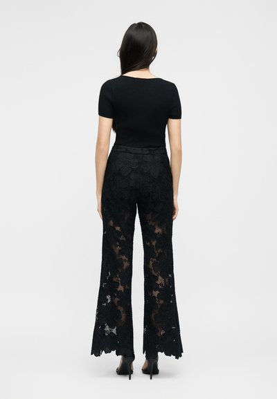 Γυναίκα στέκεται πλάτη, φορώντας εφαρμοστή μαύρη κοντομάνικη μπλούζα και ψηλόμεσα μαύρα δαντελένια παντελόνια με φαρδιά πόδια και τακούνια.