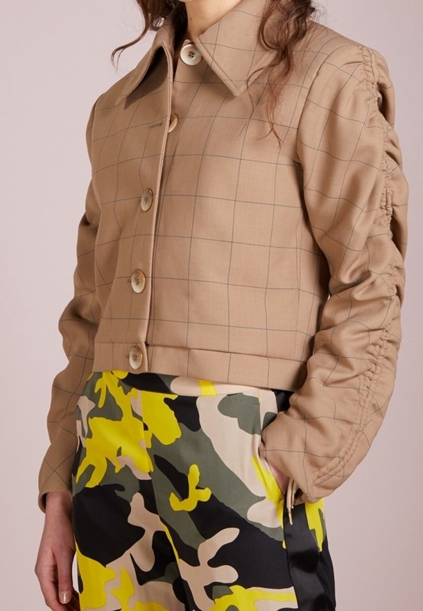 Veste à carreaux marron clair avec manches froncées associée à un pantalon taille haute avec un motif camouflage jaune, noir, blanc et vert.