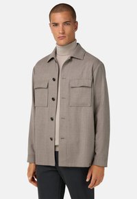 Šedá saka s knoflíky vyrobená z texturované tkaniny, s dvěma náprsními kapsami a límečkem, nošena přes světlý rolák.