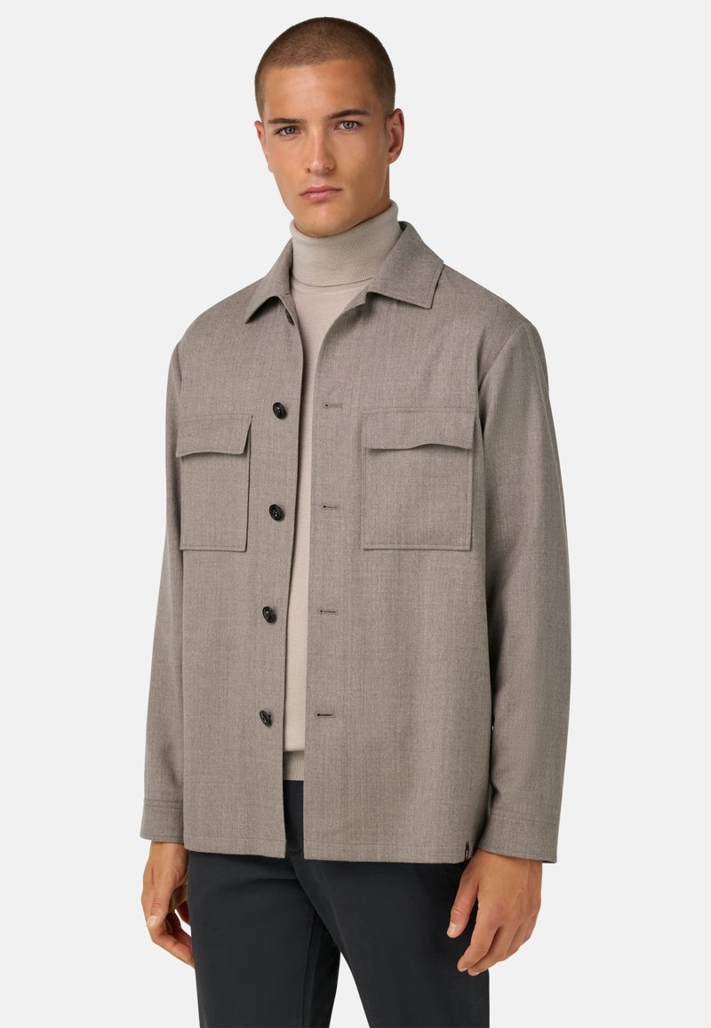 Šedá saka s knoflíky vyrobená z texturované tkaniny, s dvěma náprsními kapsami a límečkem, nošena přes světlý rolák.