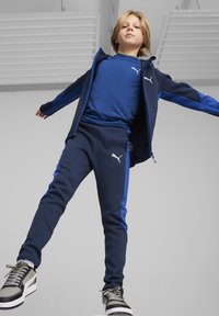 Chándal azul marino con acentos en azul claro, que cuenta con una chaqueta con cremallera y pantalones a juego. El material parece suave; incluye un logotipo en el pecho y en los pantalones.
