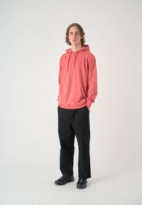 Pfirsichfarbene Kapuzenjacke mit Fronttasche und Kordelzug, kombiniert mit schwarzen weit geschnittenen Hosen und schwarzen Sneakers. Schlichtes, ungemustertes Design.