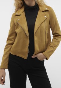 Mosterdgele jas van imitatiesuède met een getailleerd ontwerp, asymmetrische ritssluiting, metalen details en lange mouwen, gecombineerd met een zwarte coltrui.