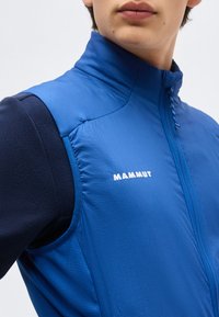 Νεαρός φοράει μπλε γιλέκο Mammut softshell πάνω από μακρυμάνικο σκούρο μπλουζάκι, κοιτώντας ελαφρώς πλάγια με το φερμουάρ στον γιακά κλειστό.