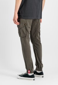 Cargo hlače v oljčni zeleni barvi, z več stranskimi žepi, elastičnimi manšeti in sproščenim krojem. Nosi jih s črnimi supergami z belimi podplati.