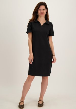 Vrouw staand, gekleed in een zwarte jurk met kraag tot op de knie en korte mouwen, en leopardprint platte schoenen, glimlachend naar de camera.