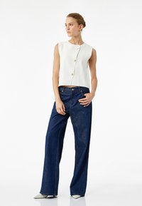 Weißes ärmelloses Oberteil mit drei Knöpfen, kombiniert mit dunkelblauen weiten Jeans. Das Oberteil hat eine glatte Textur, und die Jeans weisen aufgestickte Details auf.