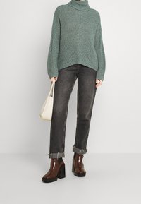 Turkusowy sweter z golfem, luźny krój, połączony z ciemnoszarymi dżinsami z postrzępionymi mankietami i brązowymi botkami na obcasie, trzymający kremową torebkę.